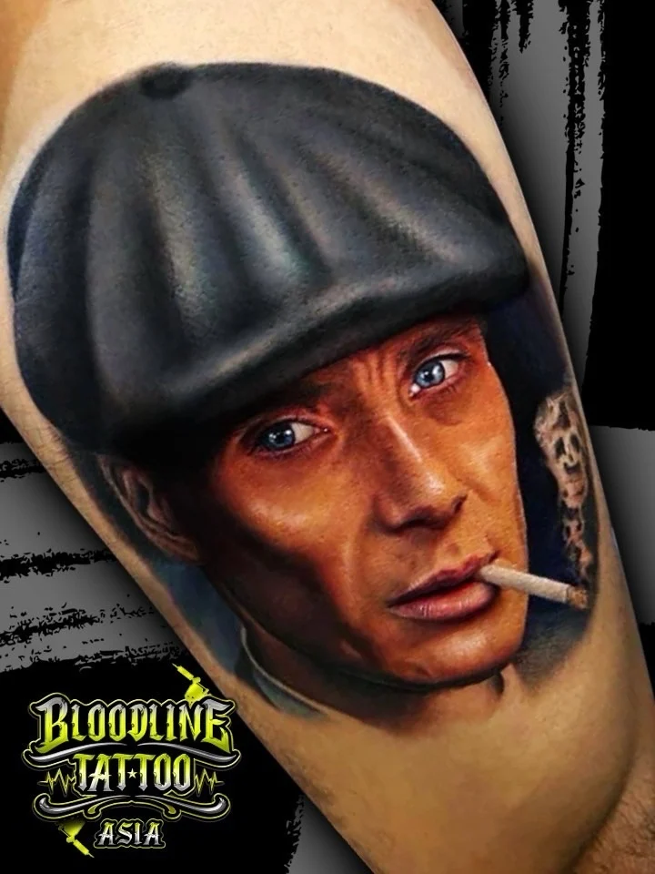 Colour portrait realism tattoo of Thomas Shelby in a flat cap with a cigarette on the forearm at Bloodline Tattoo Bangkok.