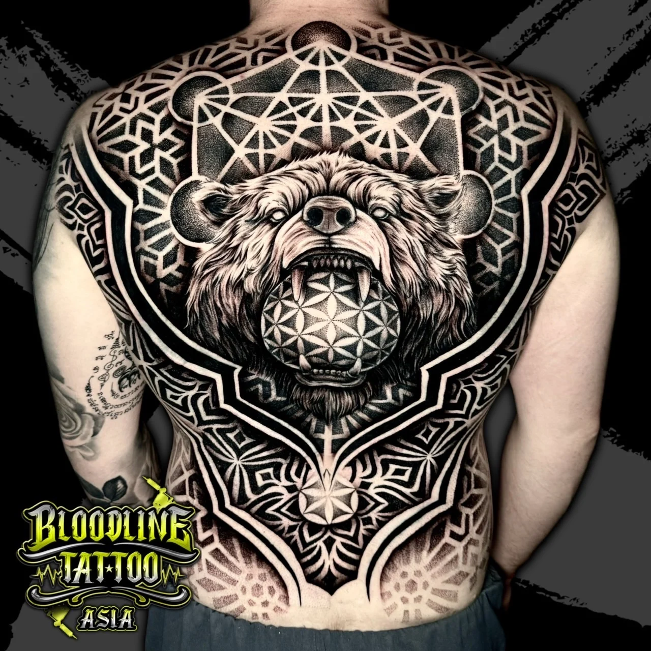 Full-back tattoo with snarling bear biting a Flower of Life sphere, Metatron's Cube, and lower mandala dotwork in black and grey, at Bloodline Tattoo Bangkok.
