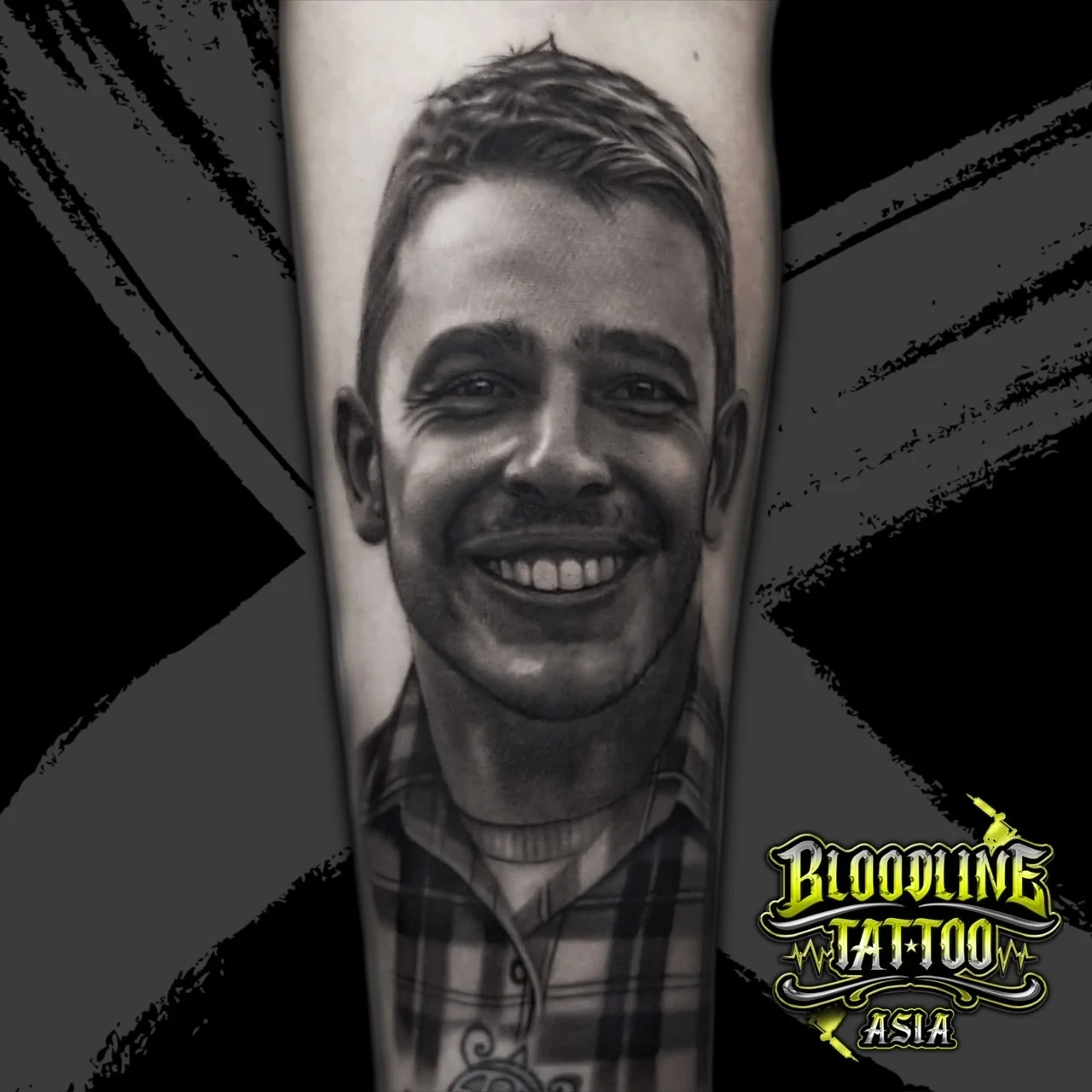 Black and grey portrait realism forearm tattoo of a smiling man in a detailed plaid button-down shirt at Bloodline Tattoo Bangkok.