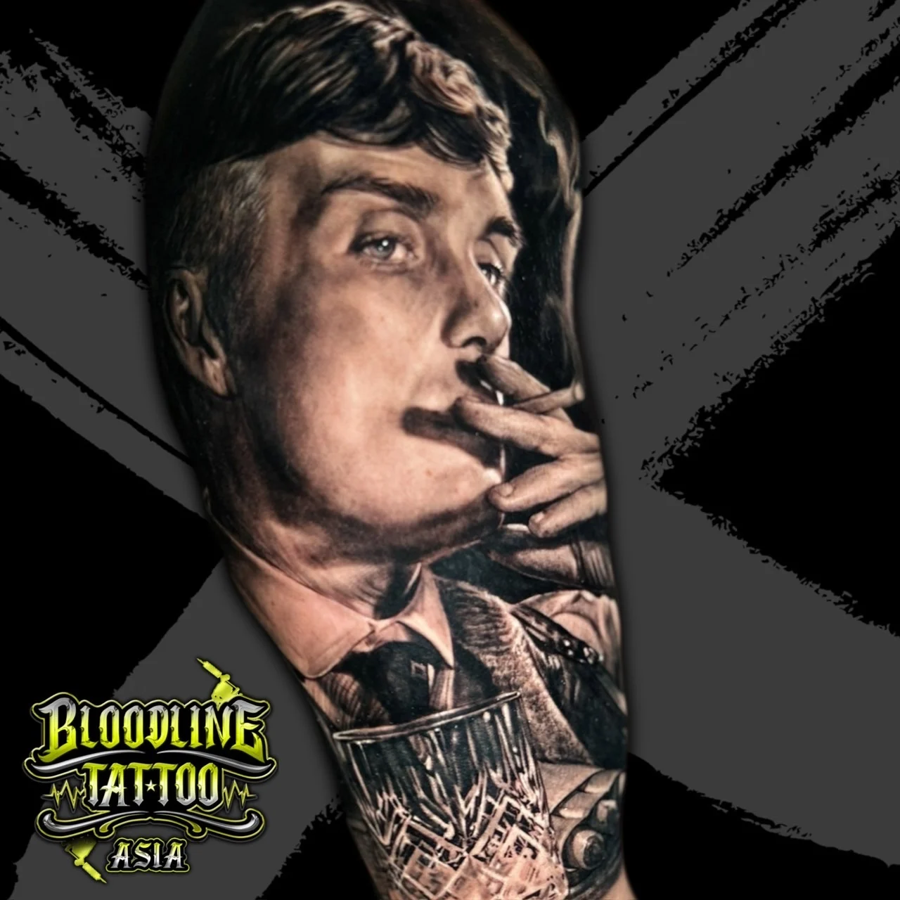 Black and grey portrait realism arm tattoo of Thomas Shelby from Peaky Blinders smoking a cigarette and holding a whiskey glass at Bloodline Tattoo Bangkok.