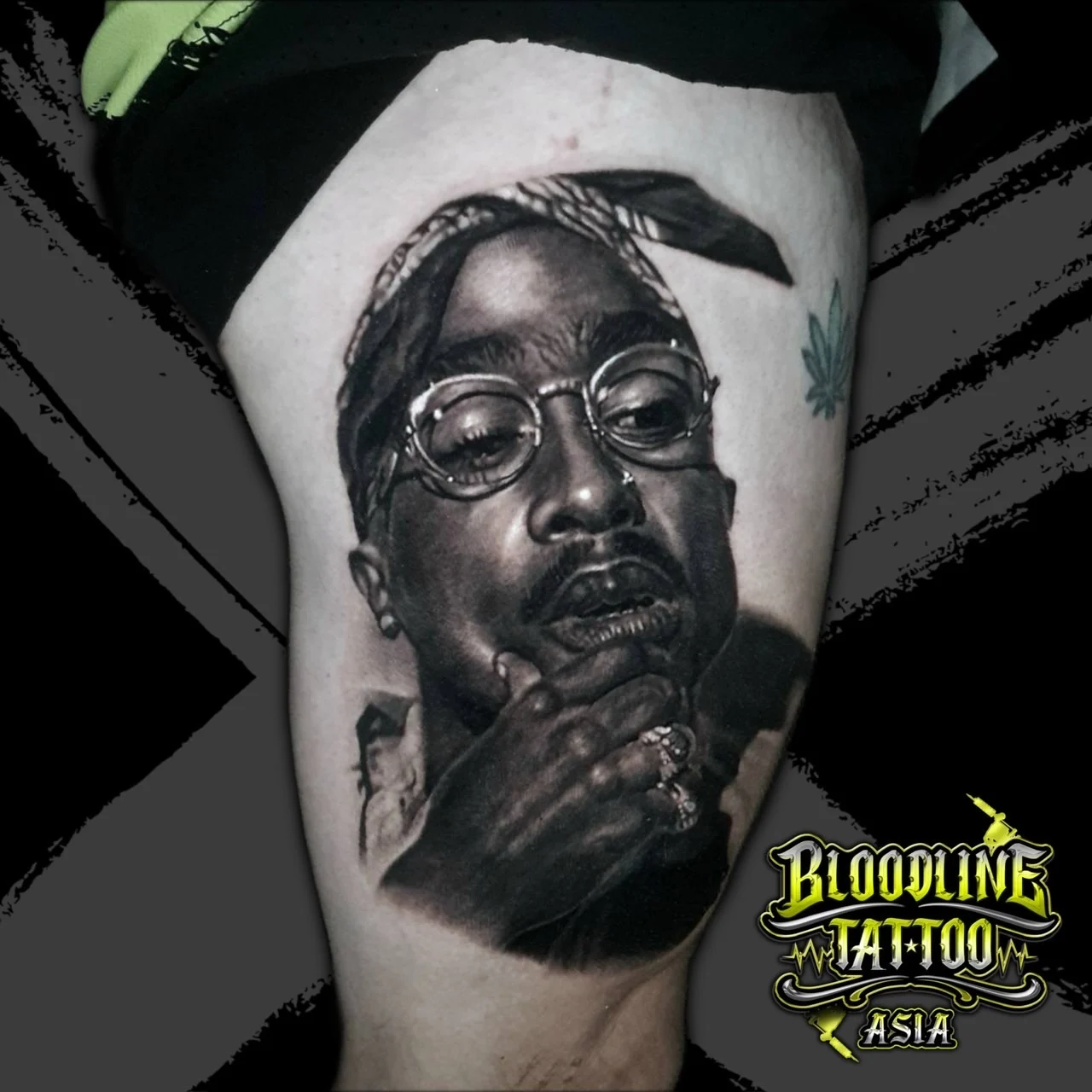 Black and grey portrait realism thigh tattoo of Tupac Shakur in bandana and round glasses with rings on his raised hand at Bloodline Tattoo Bangkok.