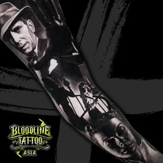 A black and grey realism sleeve with classic film-noir scenes including a fedora-clad man, shadowed figures on a staircase, and a somber woman's portrait, at Bloodline Tattoo Bangkok.