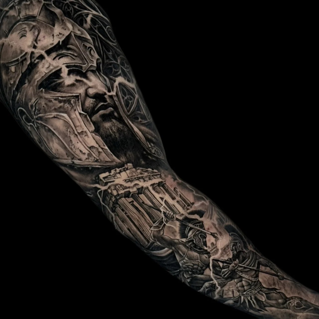 A black and grey realism full sleeve of ancient Greece with a cracked Spartan helmet portrait, a lightning-struck temple, and battling warriors on the forearm, at Bloodline Tattoo Bangkok.